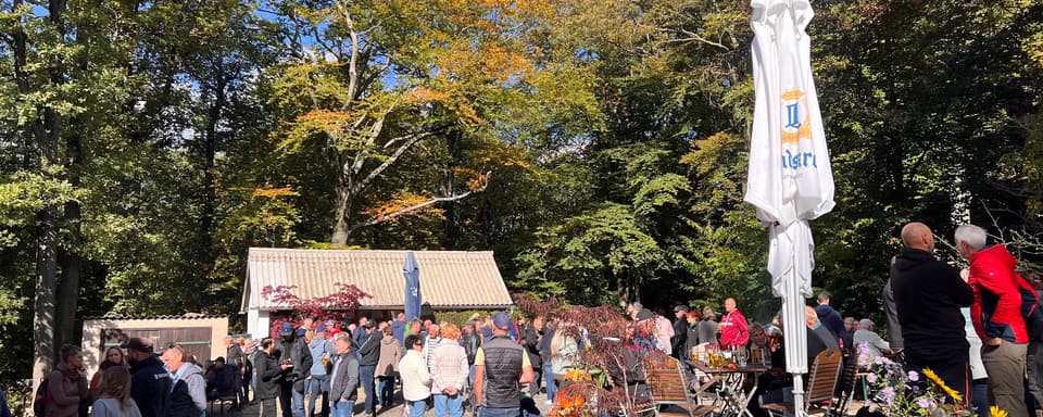 Festplatz an der Hochsteinbaude: Besucher zwischen Bierbänken und Sonnenschirmen, ringsum herbstlicher Laubwald unter blauem Himmel